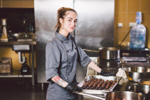 subject profession and cooking pastry. young caucasian woman with tattoo of pastry chef in kitchen of restaurant preparing round chocolate candies handmade truffle in black gloves and uniform